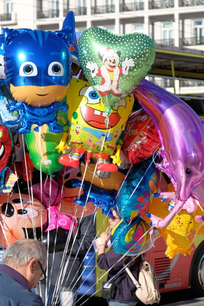 Colorful character balloons including blue superhero and green heart shapes tied to a red vehicle in an urban street setting
