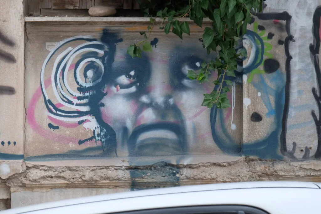 Street art mural featuring stylized face with spiral designs and pink accents on weathered concrete wall