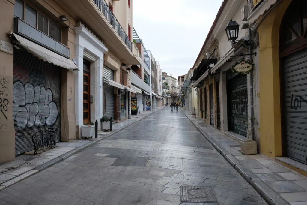 Empty narrow pedestrian street with shuttered storefronts, white and yellow building facades, and gray stone pavement in an urban European setting.