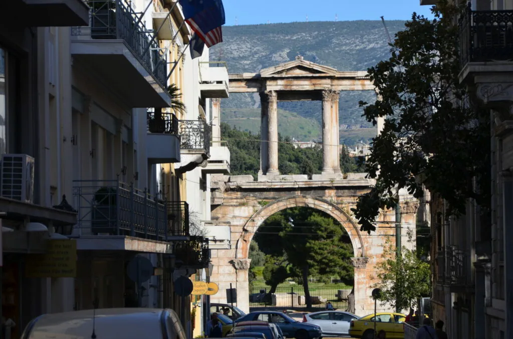Ancient stone archway with Corinthian columns framing a mountain landscape, viewed through modern city street with parked cars and residential buildings