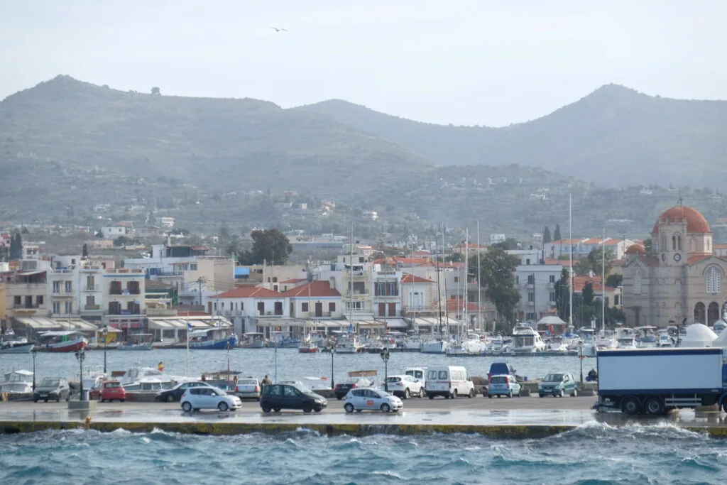 Coastal Mediterranean town with whitewashed buildings, red-roofed church, harbor with boats, and mountains in background