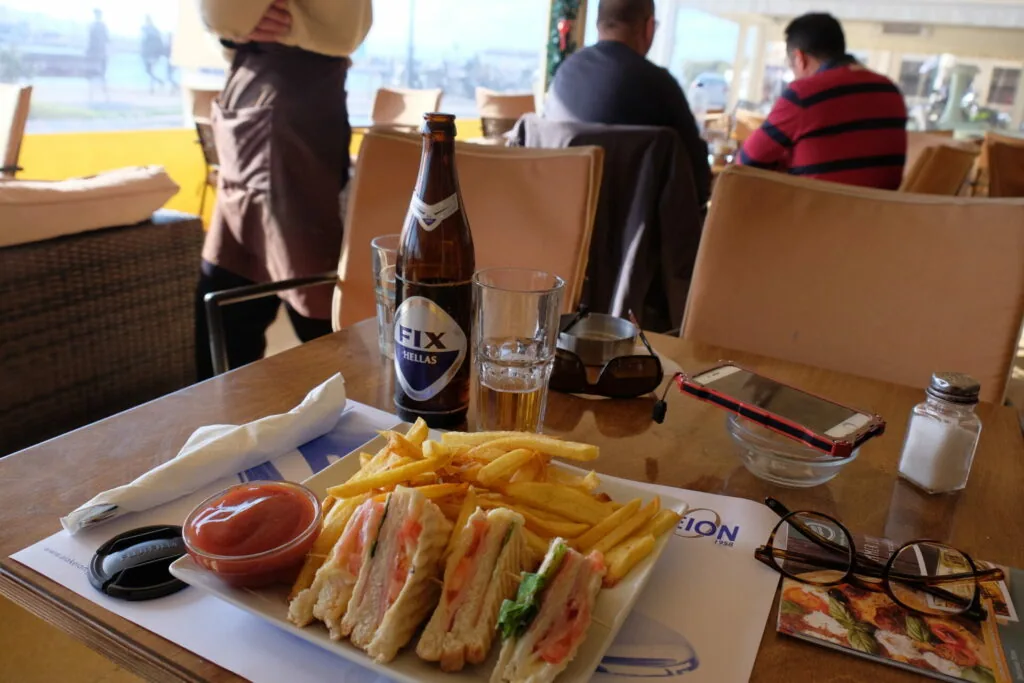 Club sandwich and fries on table with beer bottle and glass, city skyline visible through windows behind