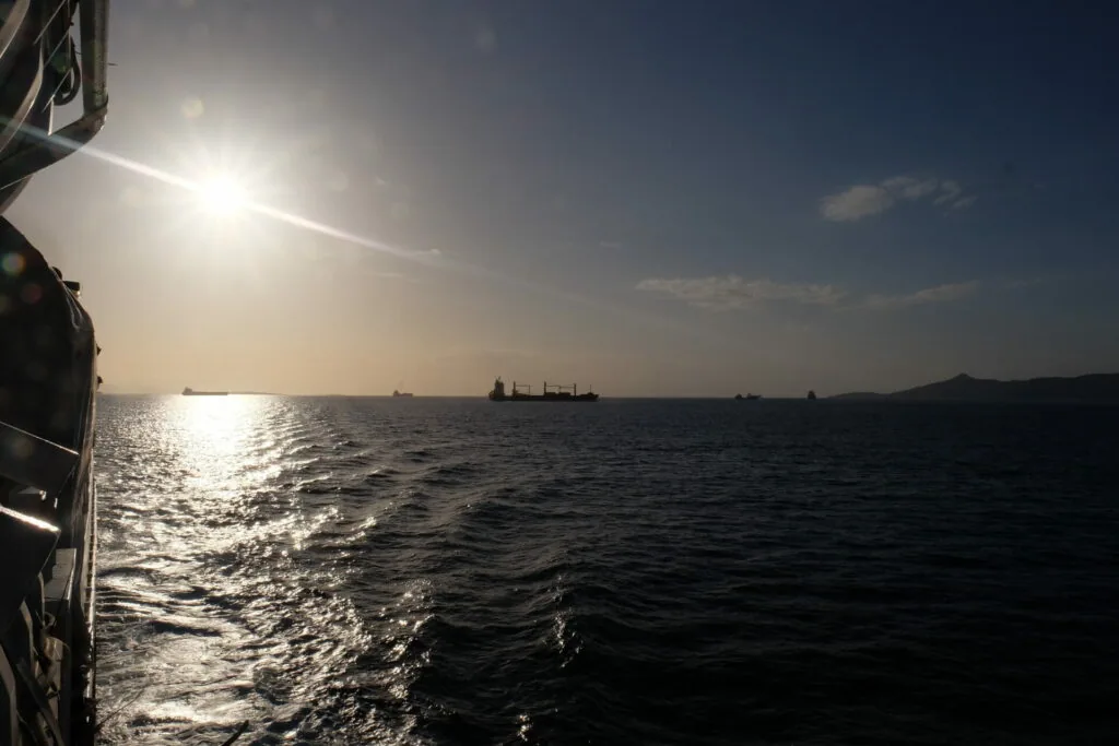 View from a boat showing calm sea waters with distant cargo ships on the horizon under bright sunlight