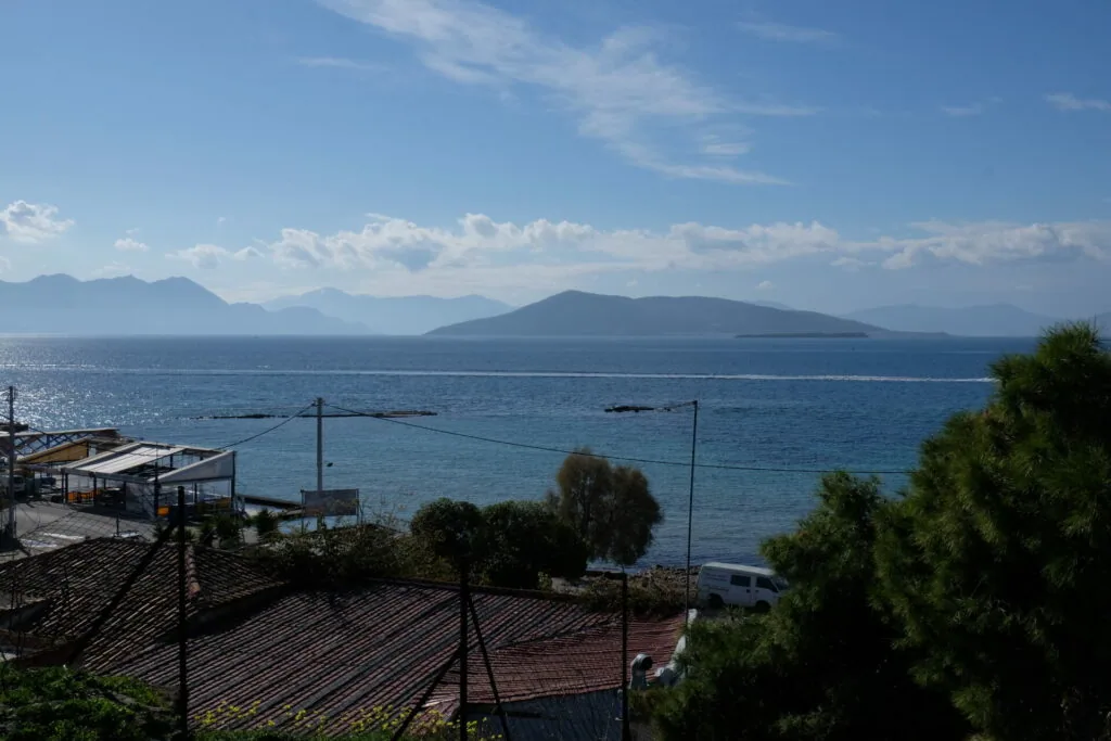 Coastal view with calm blue sea, distant mountains, waterfront buildings, and vegetation in foreground under partly cloudy sky