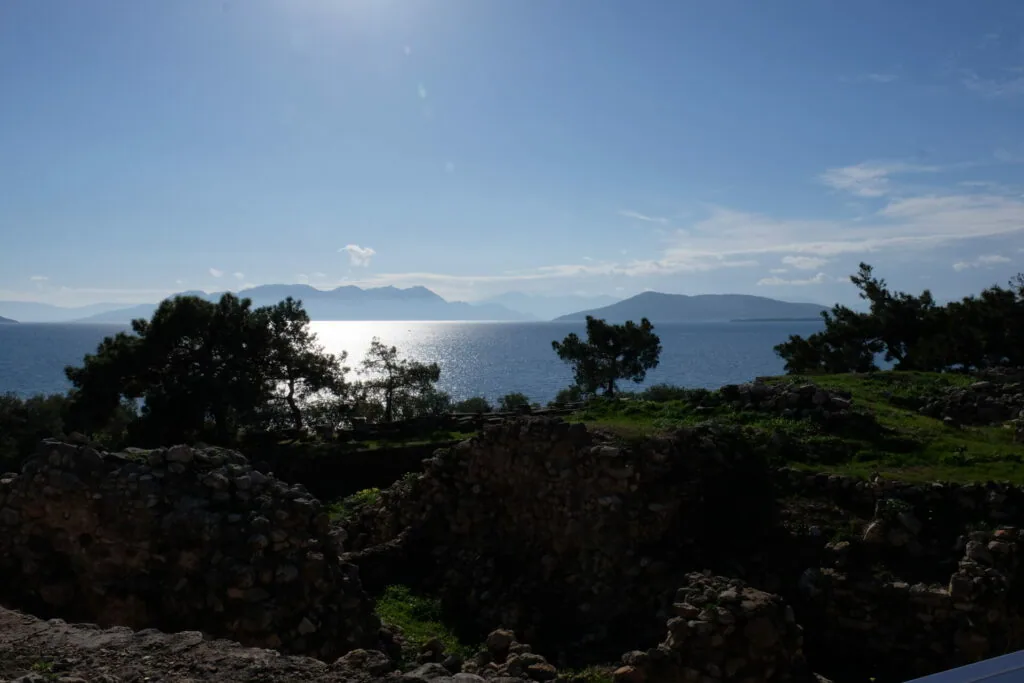 Rocky coastal landscape with scattered trees overlooking a calm blue sea with distant mountains under clear sky