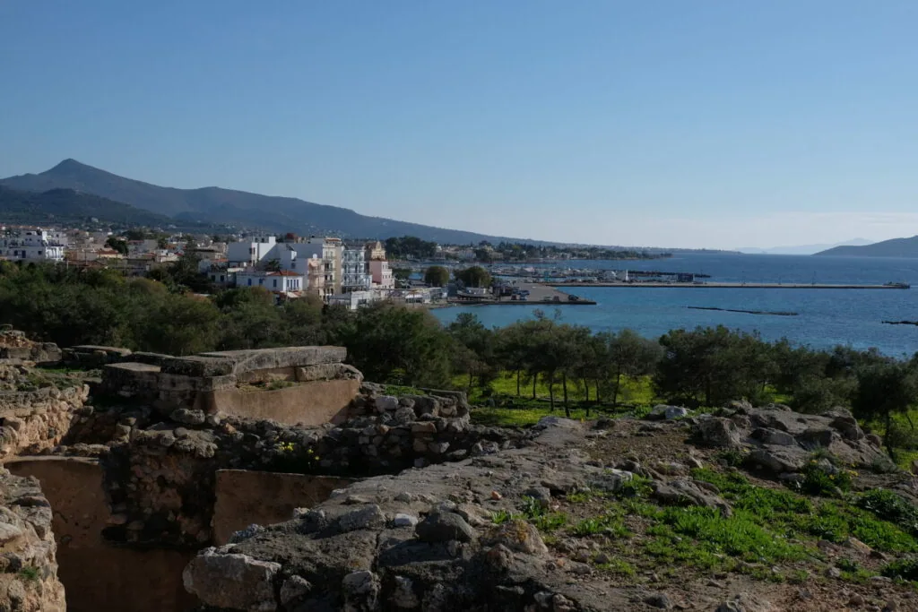 Mediterranean coastal town with white buildings, blue sea, mountains, and ancient stone ruins in foreground