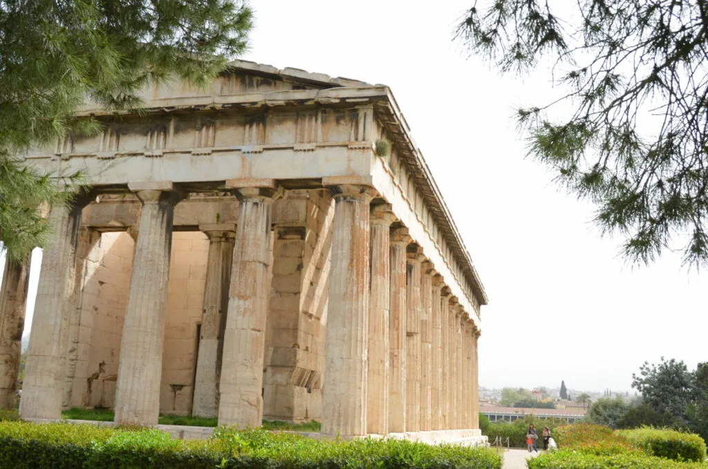 Ancient Greek temple with Doric columns, featuring fluted stone pillars and entablature, surrounded by green hedges and cypress trees.