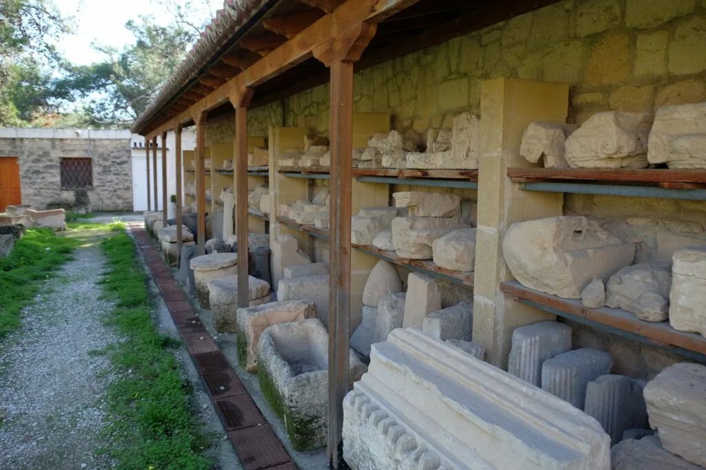 Covered storage structure with wooden shelves holding carved stone architectural fragments and sculptural pieces