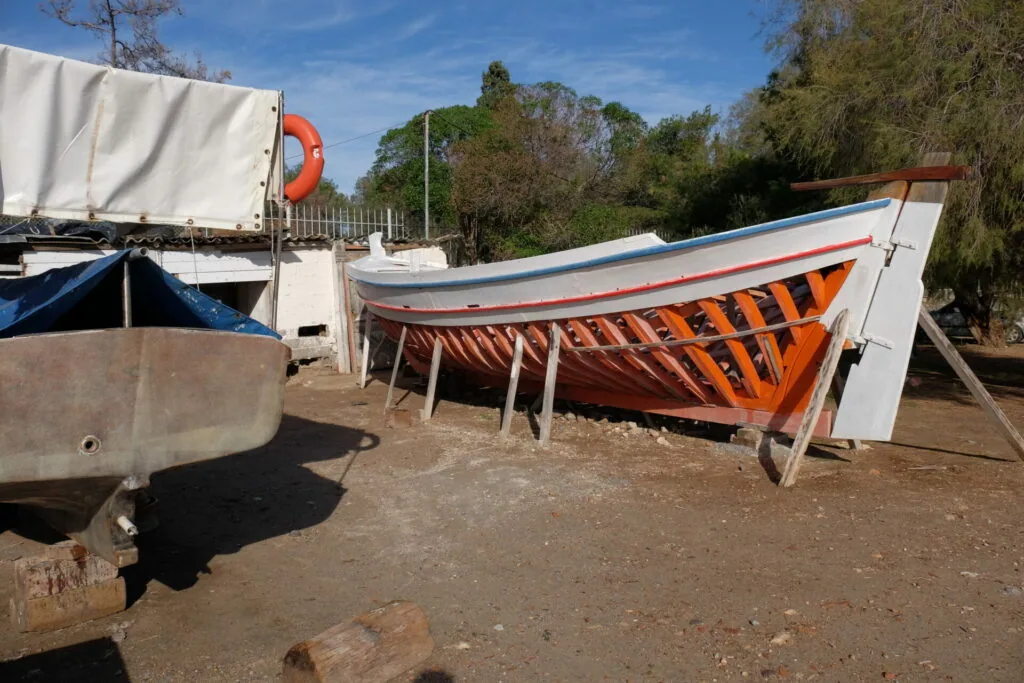 Wooden fishing boats with orange and white hulls stored on dry land, with trees and equipment in background
