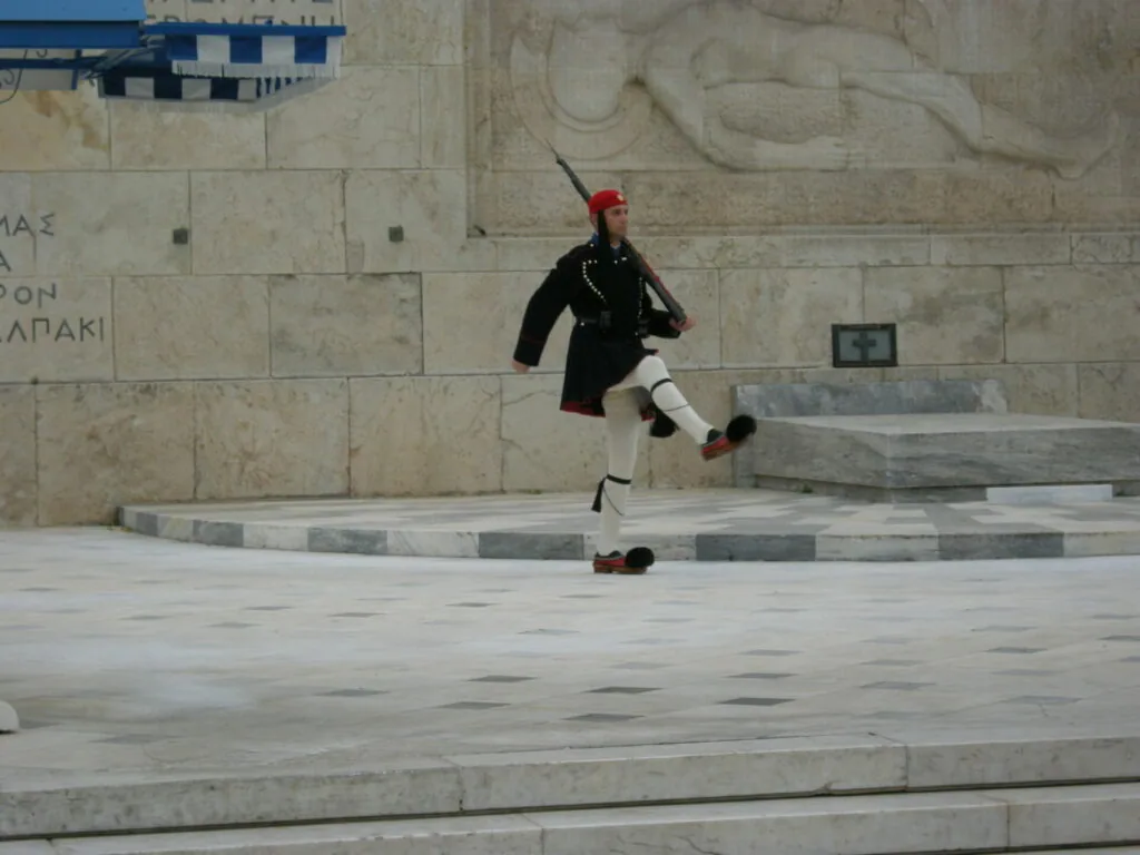 Guard in red cap and black uniform with white leg guards jumping over striped concrete barrier