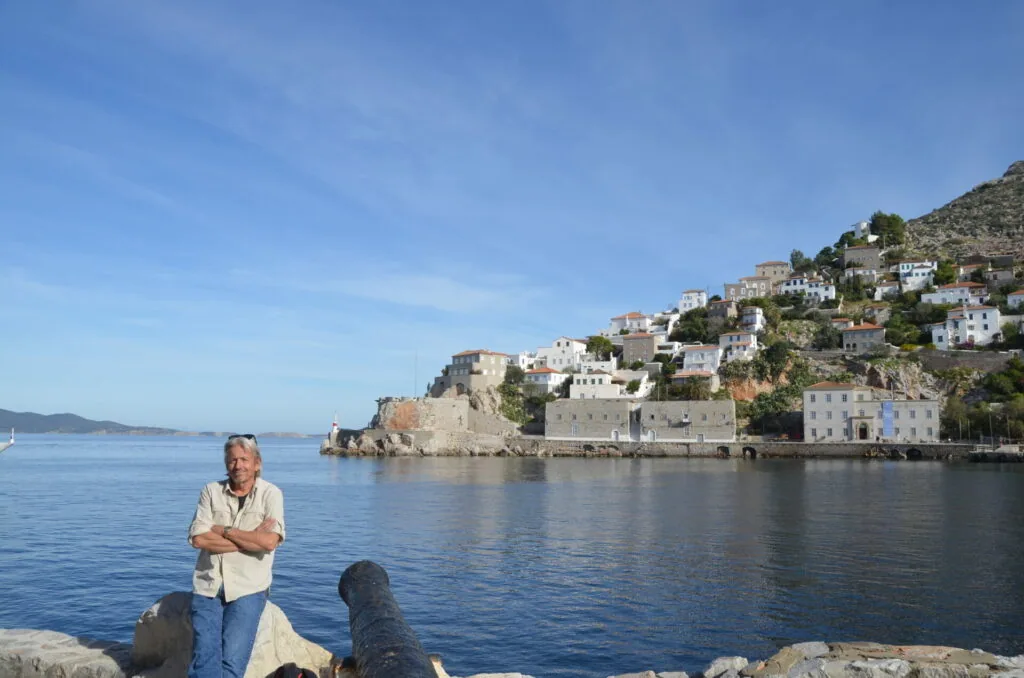 Man standing on rocky shore facing hillside village with whitewashed buildings cascading down rocky slope to calm blue water