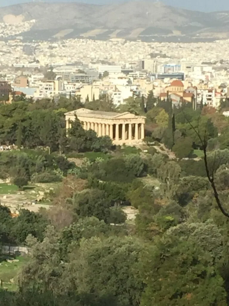 Ancient Greek temple with Doric columns surrounded by green vegetation overlooking dense urban cityscape and distant mountains