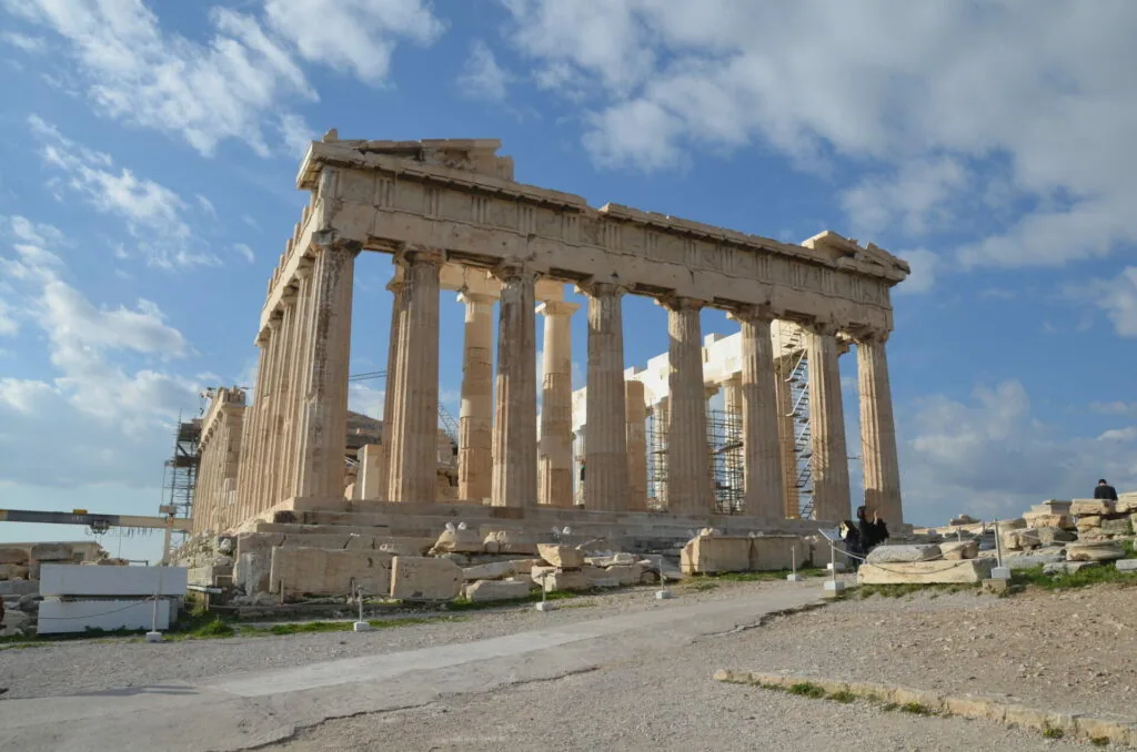 Ancient Greek temple with Doric columns and partially intact stone structure against blue sky with clouds