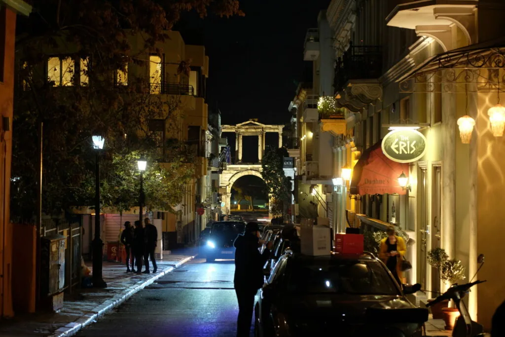 Nighttime street scene with illuminated neoclassical buildings, parked cars, pedestrians, and glowing storefronts