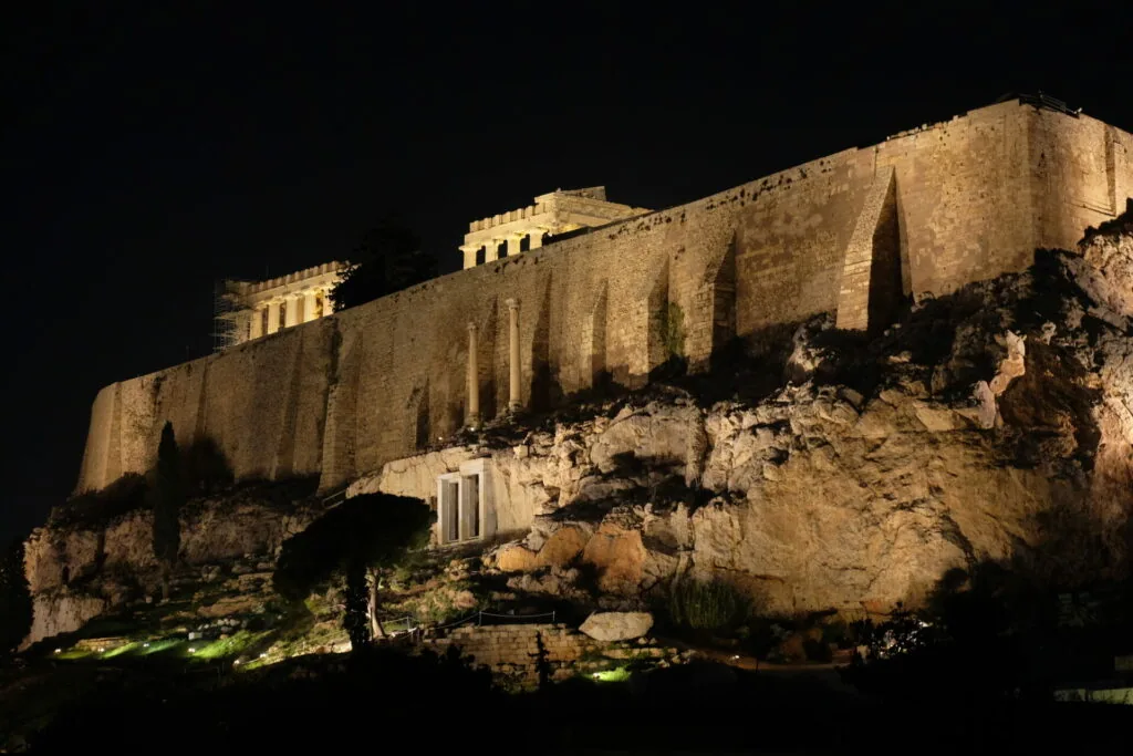 Ancient stone fortress with columned structures illuminated at night on rocky hilltop against dark sky