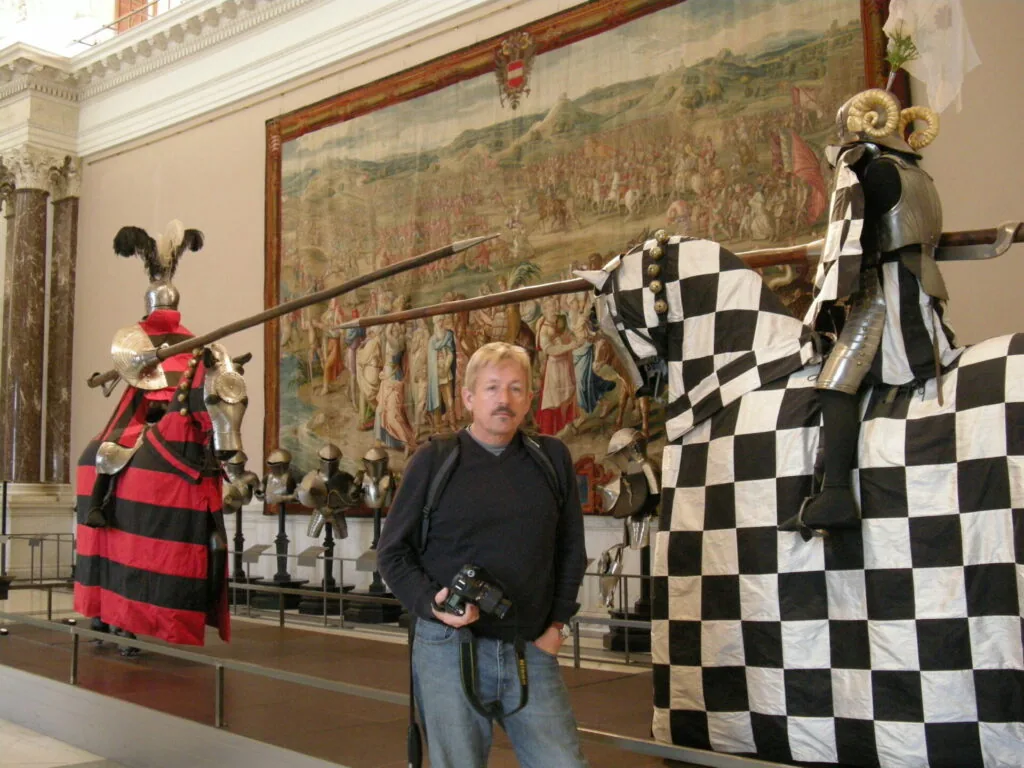 Man holding camera standing between medieval suit armor displays in museum gallery with large Renaissance fresco on wall