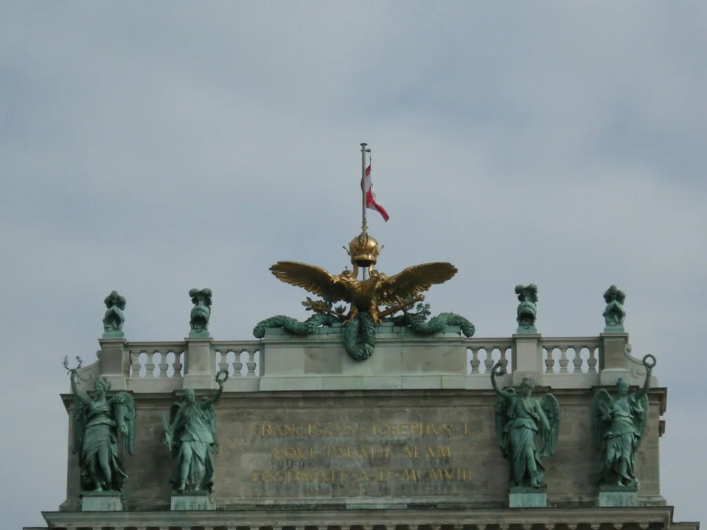 Ornate building roofline with golden eagle finial, green patina statues, and red and white flag against cloudy sky