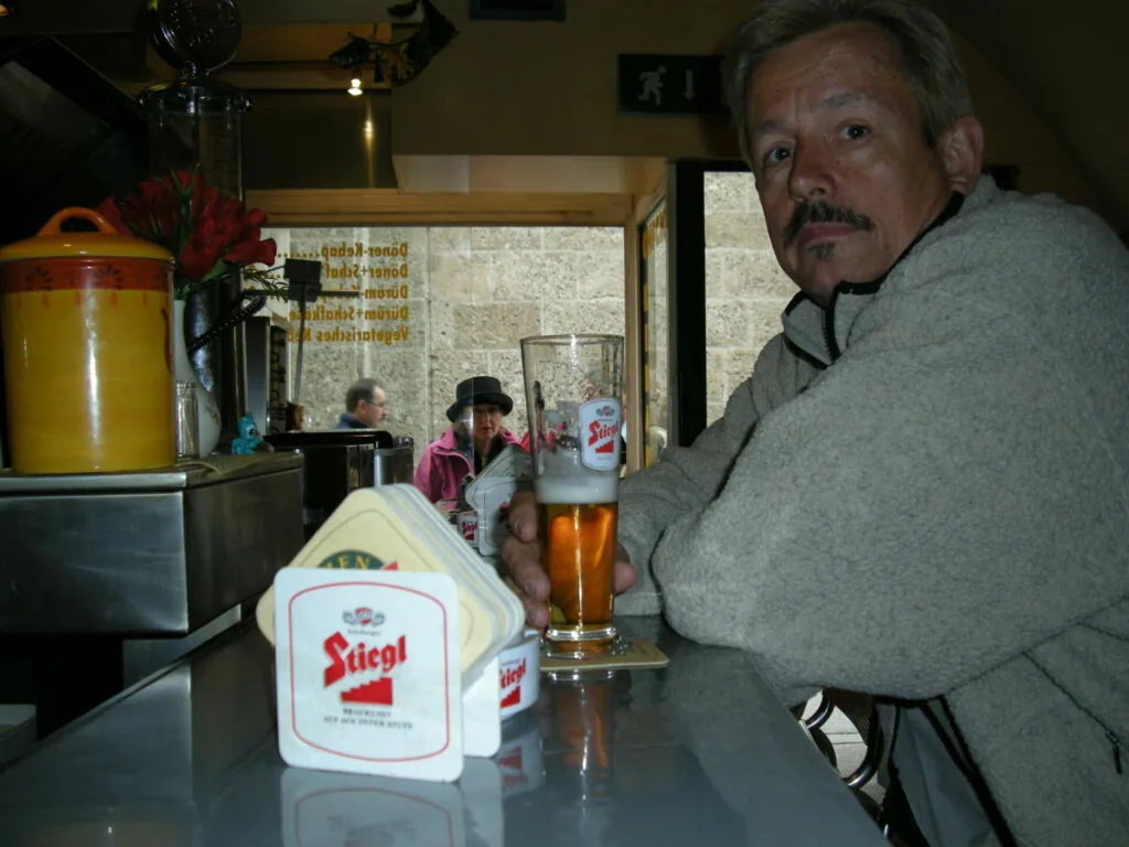Man in gray sweater holding beer glass at bar counter with beer coasters and flowers visible