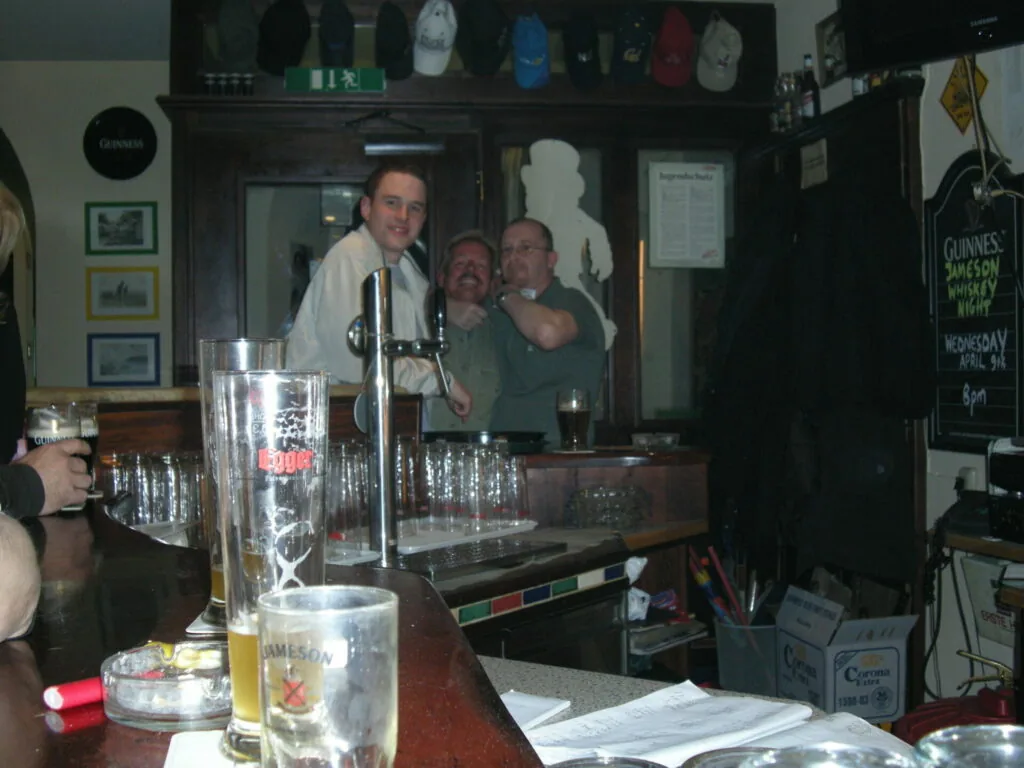 Three men behind a pub bar counter with beer glasses and bottles in foreground