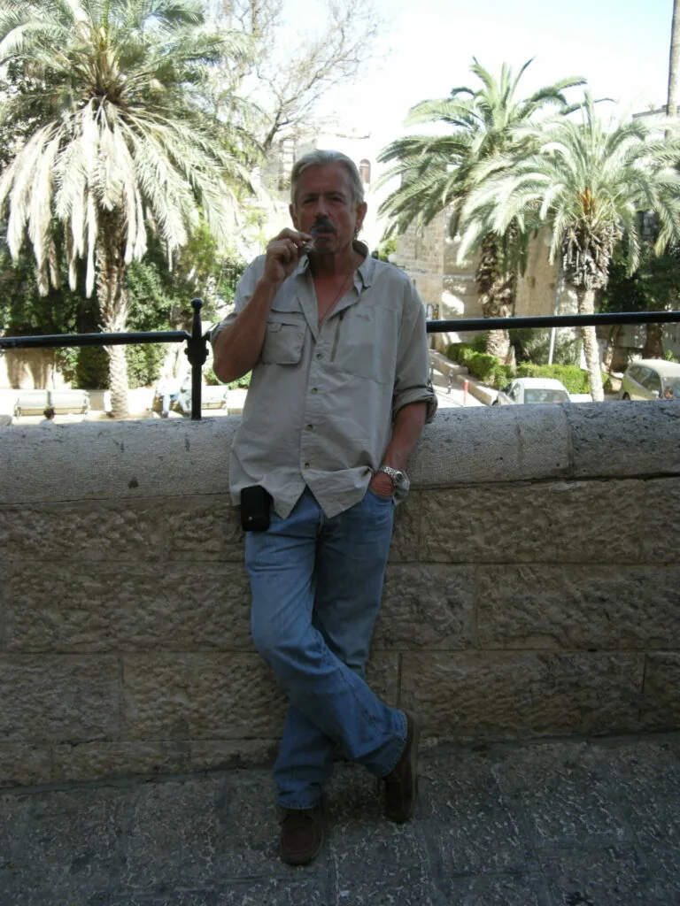 Man in light blue shirt leaning against stone wall with palm trees and historic buildings in background