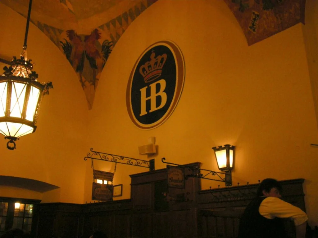 Interior of a beer hall with golden-yellow walls, hanging lamps, and a large oval logo with crown and HB initials displayed prominently