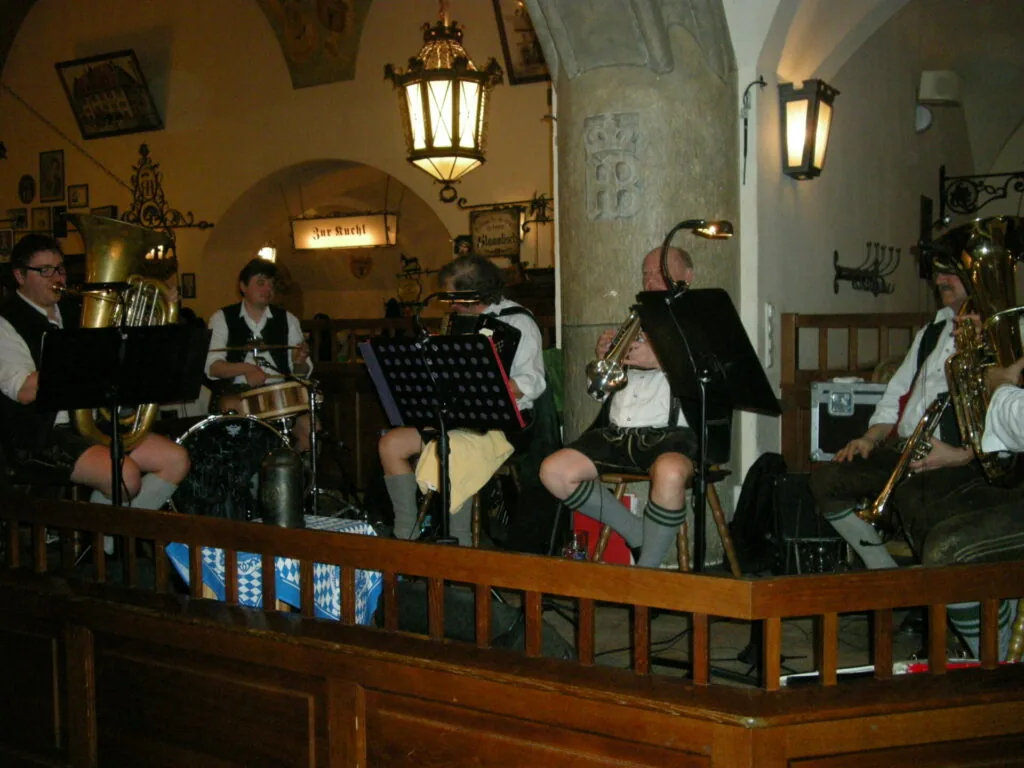 Six musicians in traditional German clothing performing with brass and wind instruments in a historic stone tavern with arched ceiling and hanging lanterns