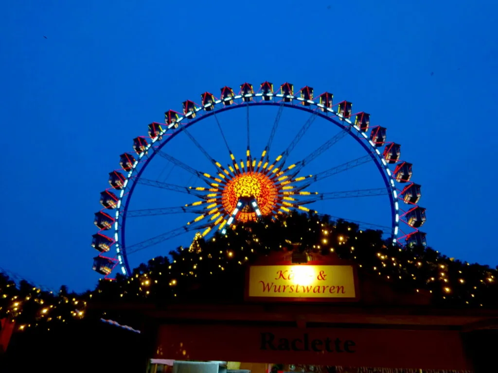 Illuminated Ferris wheel at dusk with lit garland-decorated market stall below against blue sky