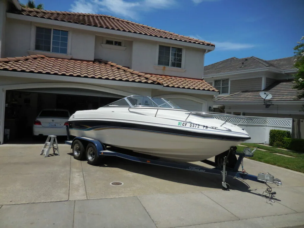 White and blue speedboat on dual-axle trailer in residential driveway with two-story house and garage in background