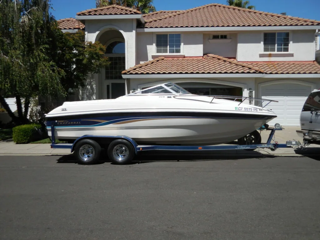 White and blue speedboat on dual-axle trailer parked in driveway in front of two-story Mediterranean-style house with tile roof