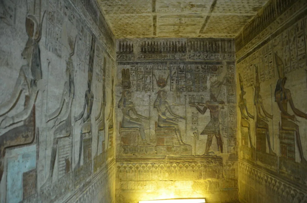 Ancient Egyptian tomb interior with hieroglyphics and painted figures on walls and ceiling, illuminated by yellow lighting.