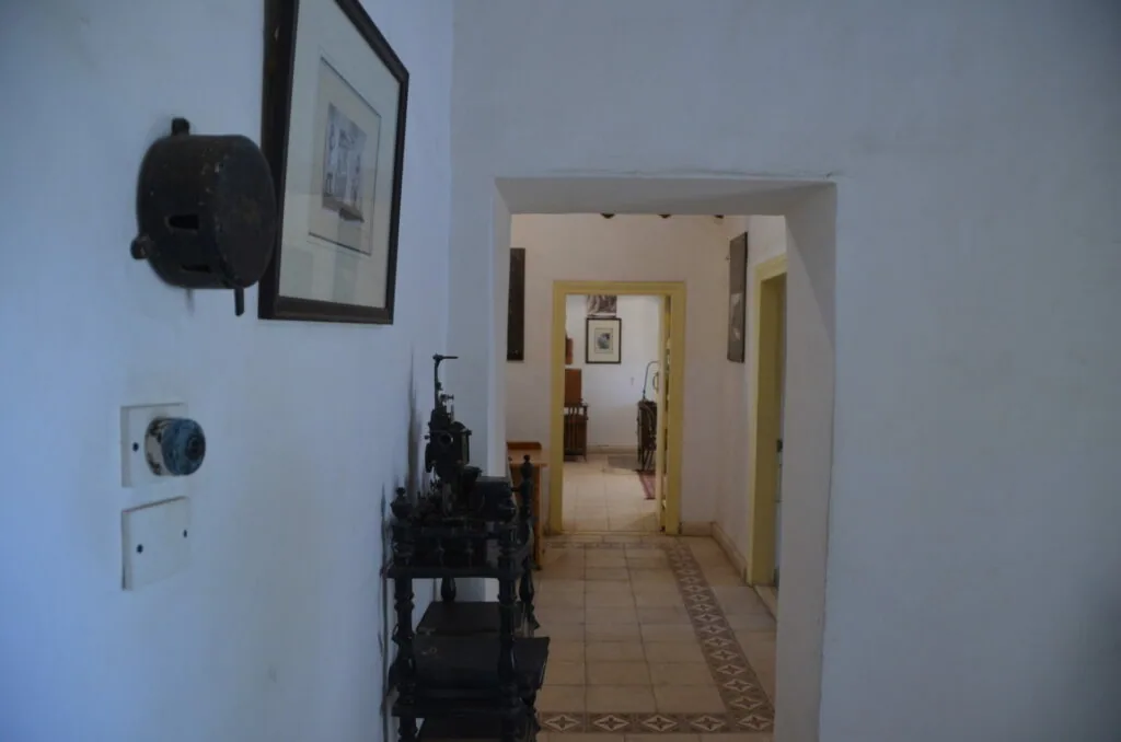 Narrow hallway with white walls, framed artwork, wall sconce, electrical outlet, and view through doorway to adjoining rooms with tile flooring