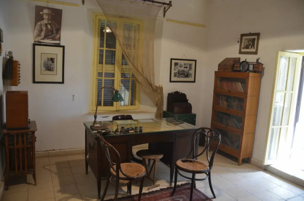 Historic study room with wooden desk, bentwood chairs, bookshelves, framed photographs on walls, and period furnishings