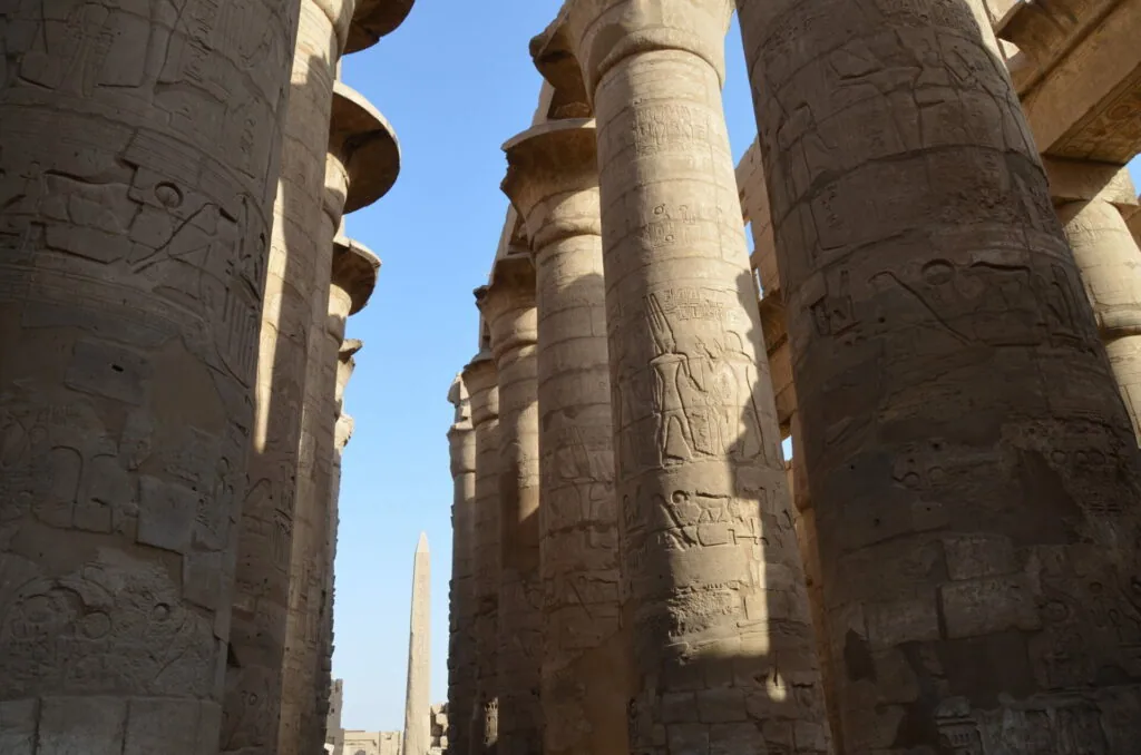 Ancient Egyptian stone columns carved with hieroglyphics and relief figures, with an obelisk visible in the background against a clear blue sky