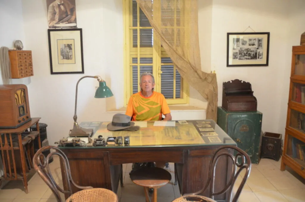 Man seated at wooden desk in vintage-style office with period furniture, framed photographs, and a green desk lamp