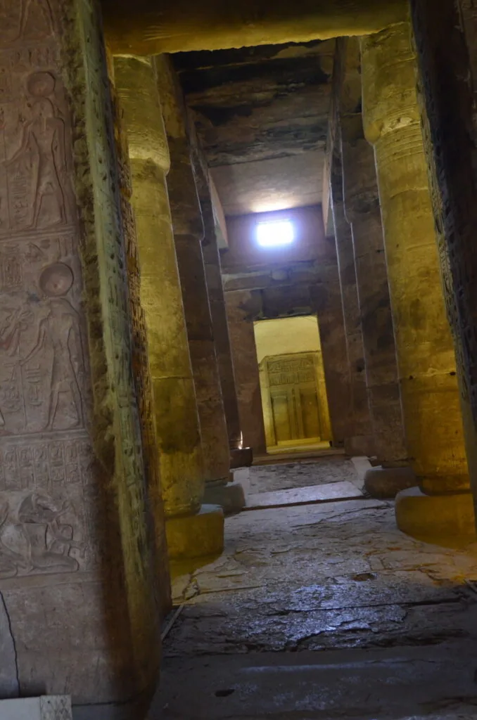 Ancient stone corridor with yellow-ochre painted walls and hieroglyphic reliefs, narrow passageway between massive columns receding into distance with blue light source visible at far end