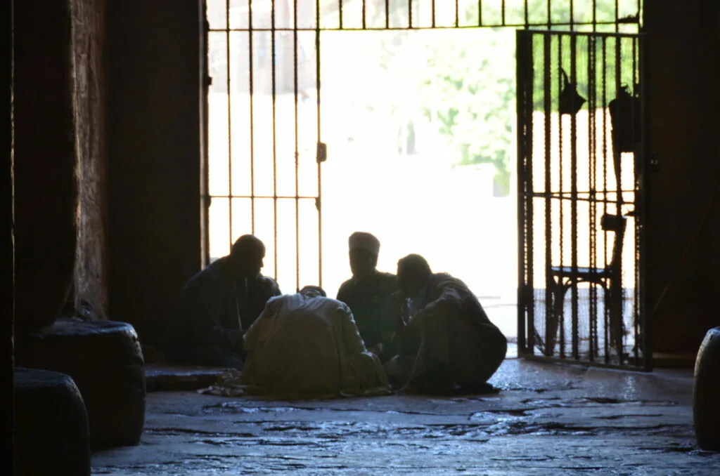 Group of people sitting in silhouette inside a dim doorway with metal gates, bright sunlight visible outside