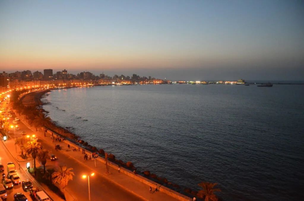 Illuminated coastal promenade at dusk with palm trees, street lights, and calm bay waters with distant ships and buildings along the shoreline