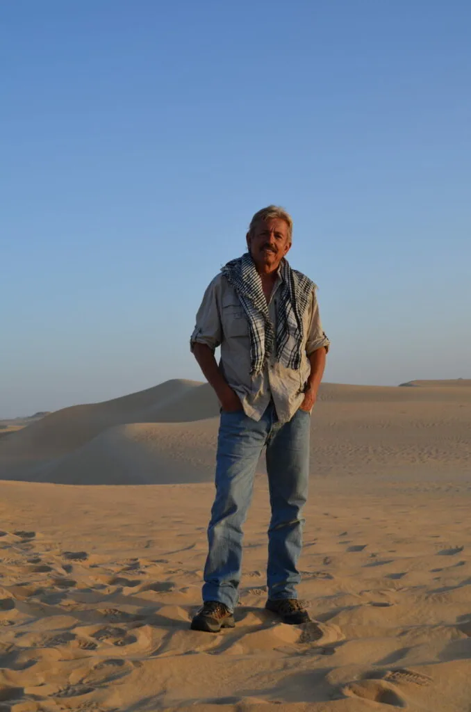 Man standing on desert sand dunes wearing a scarf and casual shirt under blue sky