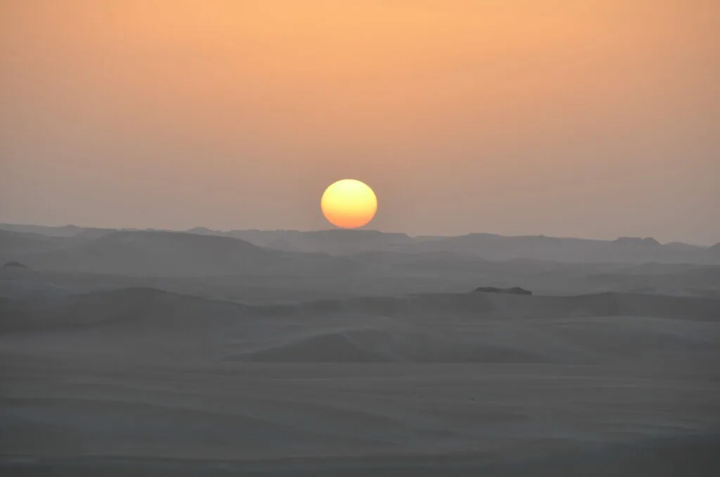 Large sun setting over layered desert hills in shades of orange and gray