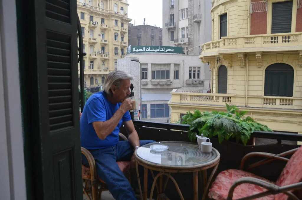 Man in blue shirt sitting on a balcony overlooking historic buildings with Arabic signage