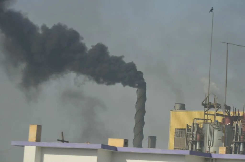 Industrial facility with tall chimney emitting thick dark smoke against gray sky