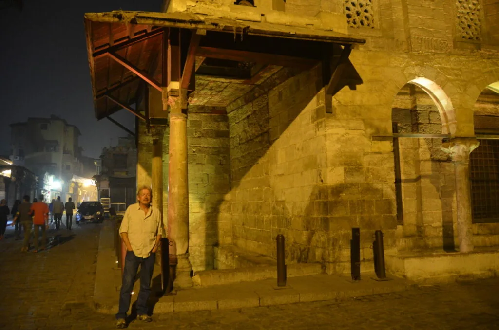 Man standing at night in front of an illuminated stone building with arched doorway and metal bollards