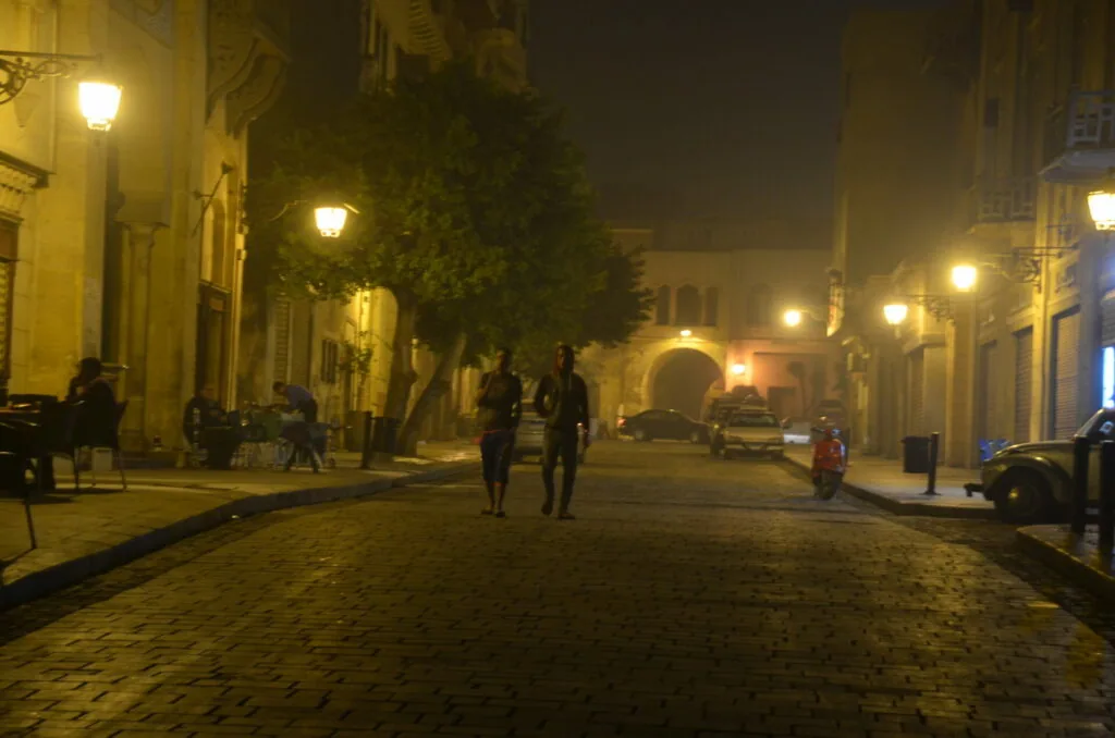 Nighttime street scene with golden street lights, colonial buildings, and pedestrians walking on a cobblestone street