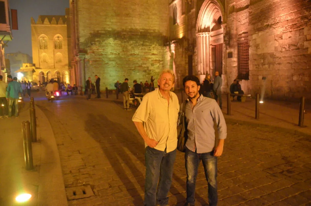 Two men posing in front of illuminated historic stone buildings and architecture at night