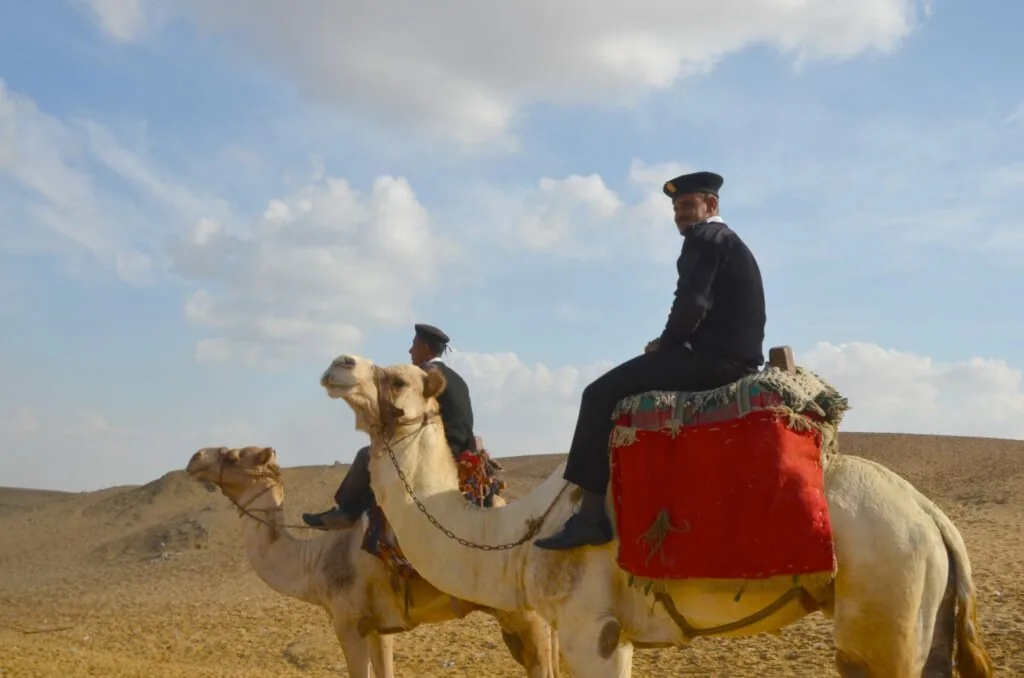 Two men in dark clothing and caps riding a camel with red saddle blanket in a sandy desert landscape