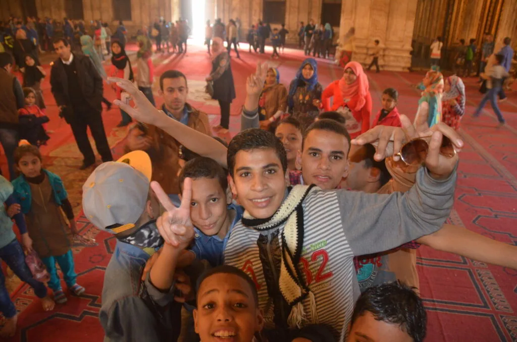 Group of children posing together inside a large red-carpeted hall with many people gathered in the background