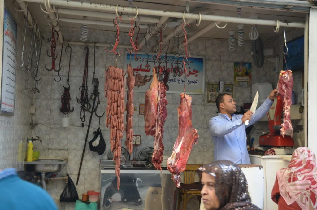 Butcher shop with hanging cuts of red meat and a man in light blue shirt holding a knife, customer visible in foreground