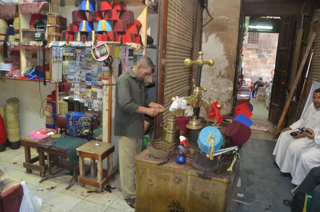 Man working at wooden table in cluttered shop with colorful hanging items and merchandise displayed on walls