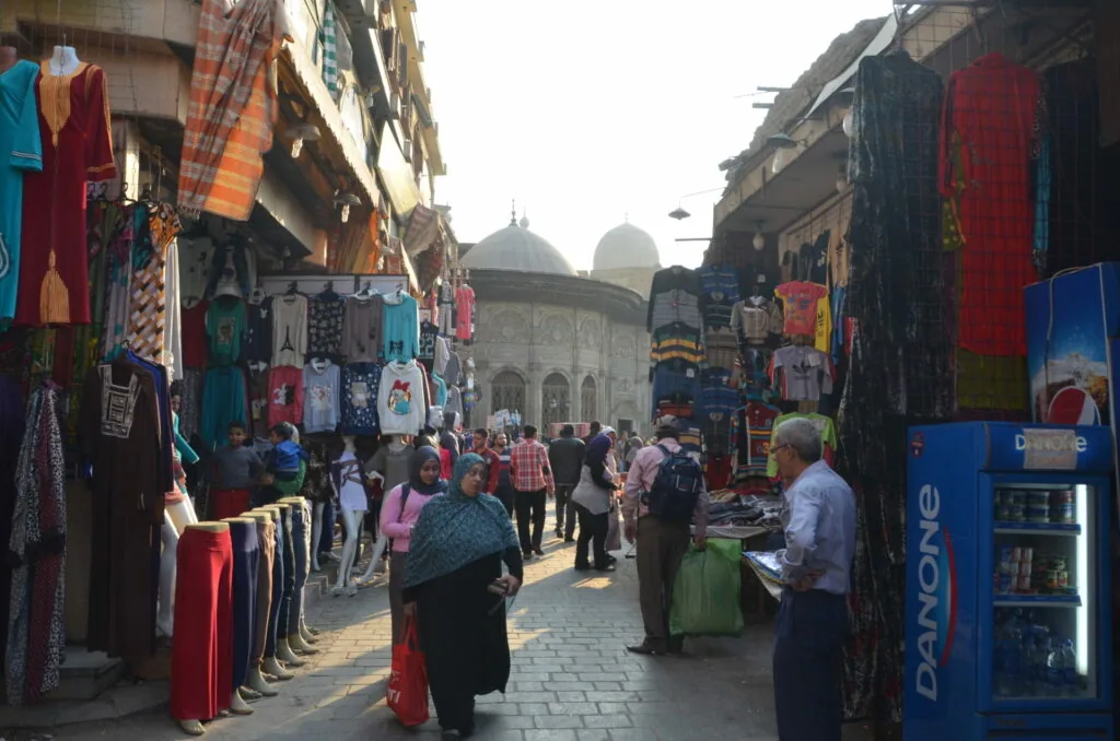 Busy marketplace street with clothing vendors on both sides, historic domed mosque visible in background, crowds of shoppers walking through narrow passage