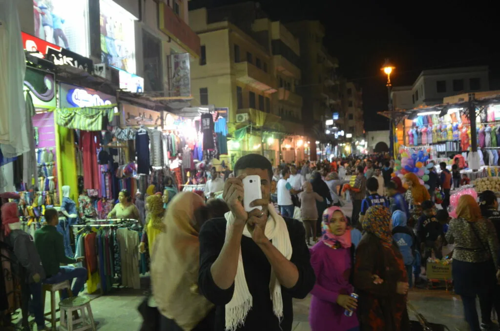 Crowded night market street with vendor stalls displaying colorful clothing and merchandise, shoppers browsing under bright lights